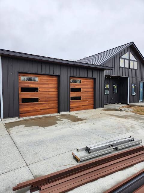 Modern dark metal building with two wood-clad garage doors and construction materials on the driveway.